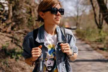 Close up portrait of stylish girl with light hair in sunglasses and denim jacket is looking away with smile in sunlight on background of forest