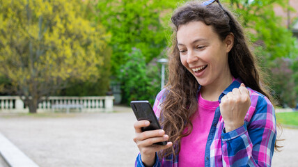 Excited young woman student doing winner gesture, looking at smartphone standing in green park...