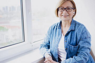 Beautiful blonde mature woman smiling looking at camera. wearing blue denim shirt. eyeglasses and wireless headset, standing near window indoor. Senior and technology. Elderly student. Copy space