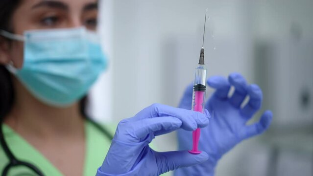 Close-up syringe with liquid drug in female Caucasian hands indoors. Blurred young woman in coronavirus face mask preparing Covid-19 vaccine injection in hospital. Slow motion