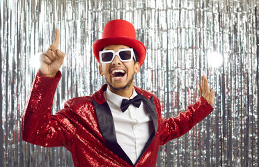 Lets get party. Positive funny young dark-skinned showman in shiny suit on silver foil background. Joyful crazy man in glasses, red hat and in suit with sequins raises his finger in sign of idea. - obrazy, fototapety, plakaty