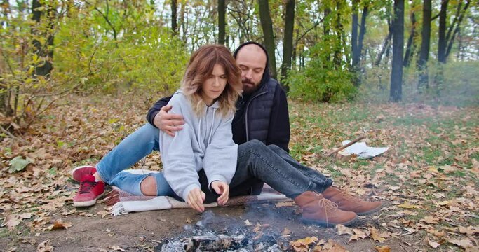 a happy girl, together with her boyfriend, sits hugging by the fire, warms her hands, on a cold autumn day. family vacation young family outdoors