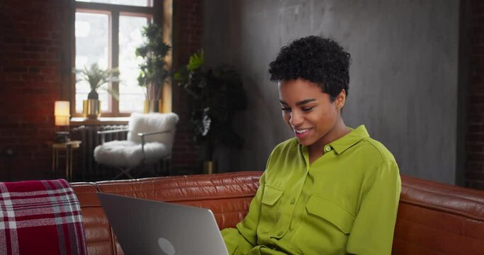 Smiling black millennial woman spend weekend at home sit on couch chat with friend in web conference virtual app using laptop. 