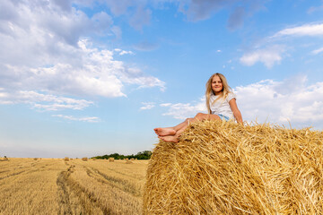 Naklejka na ściany i meble Portrait of cute little blond beautiful adorable cheerful caucasian kid girl enjoy sitting on hay stack or bale on harvested wheat field warm summer evening. Scenic rural country landscape background