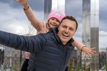 Happy father carrying piggyback ride to his daughter outdoors. Cheerful girl piggyback riding dad...
