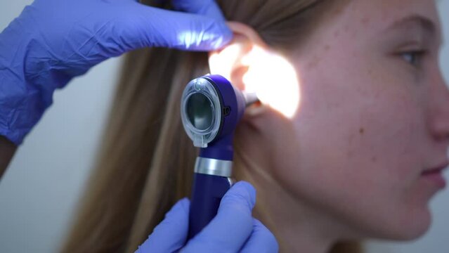 Close-up side view ear of female teenage patient with rack focus to otoscope in doctor hand. Unrecognizable otolaryngologist checking auditory canal and tympanic membrane in hospital indoors