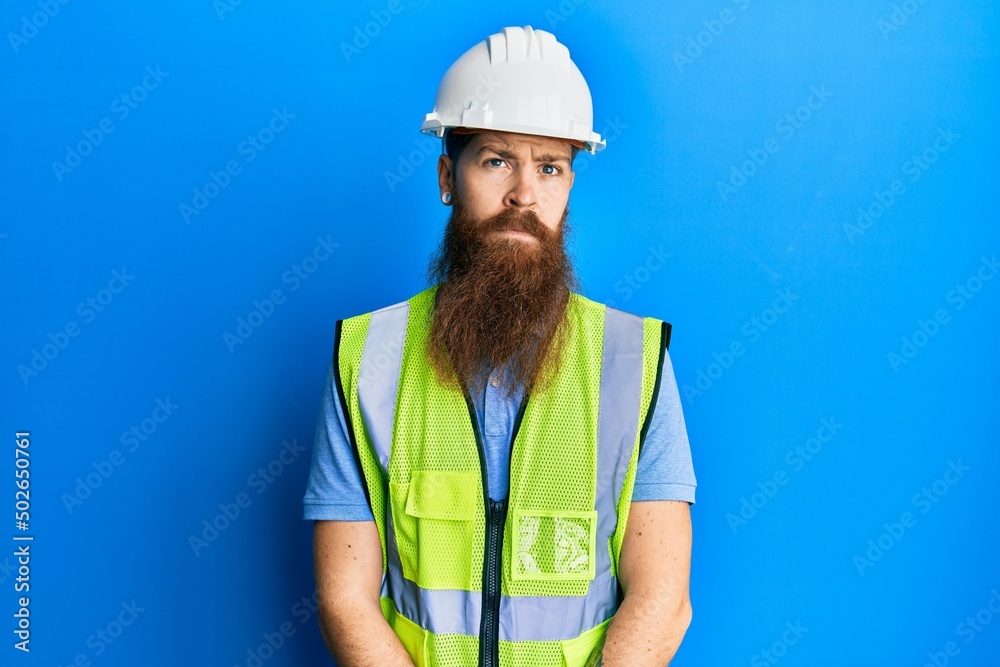 Poster redhead man with long beard wearing safety helmet and reflective jacket relaxed with serious express