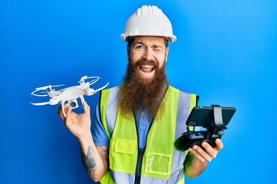 Redhead man with long beard wearing safety hard using drone with remote control winking looking at the camera with sexy expression, cheerful and happy face.