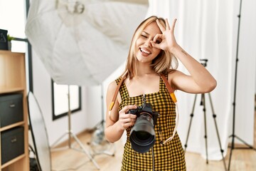 Fototapeta na wymiar Young caucasian photographer girl holding professional camera at photography studio smiling happy doing ok sign with hand on eye looking through fingers