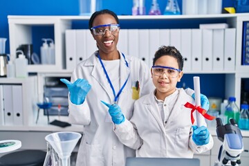 Mother and son at scientist laboratory pointing thumb up to the side smiling happy with open mouth