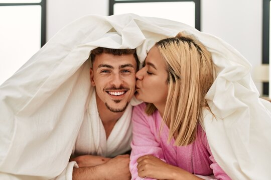 Young caucasian couple covering with bedsheet and smiling happy lying on the bed at home.