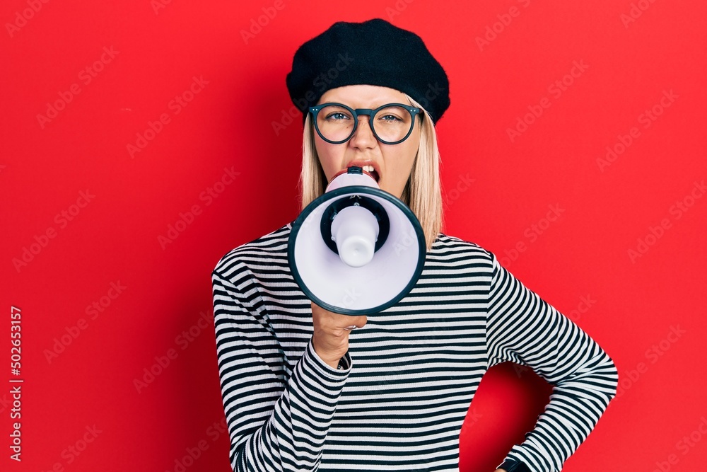 Wall mural Caucasian blonde woman wearing glasses and beret screaming with megaphone over red background