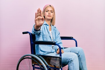 Beautiful blonde woman sitting on wheelchair doing stop sing with palm of the hand. warning...