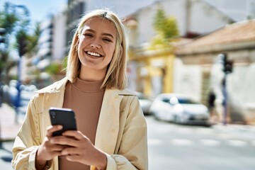 Young blonde girl smiling happy using smartphone at the city.