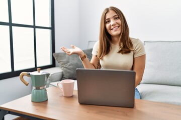 Young brunette woman using laptop at home drinking a cup of coffee smiling cheerful presenting and pointing with palm of hand looking at the camera.