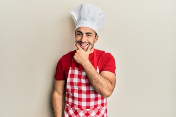 Young hispanic man wearing baker uniform looking confident at the camera smiling with crossed arms and hand raised on chin. thinking positive.