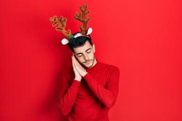 Young hispanic man wearing cute christmas reindeer horns sleeping tired dreaming and posing with hands together while smiling with closed eyes.