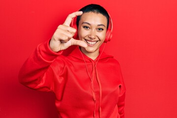 Beautiful hispanic woman with short hair listening to music using headphones smiling and confident gesturing with hand doing small size sign with fingers looking and the camera. measure concept.