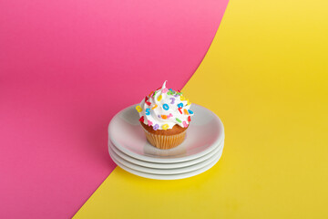 Cupcake with colorful sprinkles on a white plate, set against a split pink and yellow background.