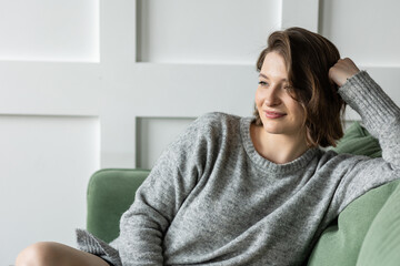 Beautiful young woman relaxed on the couch in the living room at home. Daylight. Female in a grey sweater filling comfortable at her house. Weekend
