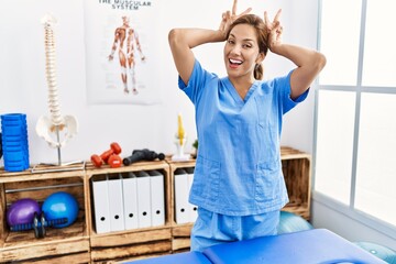 Middle age hispanic physiotherapist woman working at pain recovery clinic posing funny and crazy with fingers on head as bunny ears, smiling cheerful