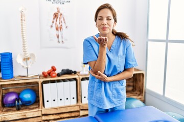 Middle age hispanic physiotherapist woman working at pain recovery clinic looking at the camera blowing a kiss with hand on air being lovely and sexy. love expression.