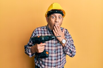 Handsome senior man with grey hair holding screwdriver wearing hardhat covering mouth with hand, shocked and afraid for mistake. surprised expression