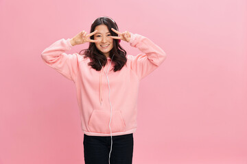 Cheerful Asian student young lady in pink hoodie sweatshirt with cute headphones show v-sign gesture posing isolated on over pink studio background. Good offer. Sound streaming platform ad concept