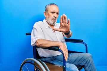 Handsome senior man with beard sitting on wheelchair doing stop sing with palm of the hand. warning...