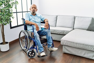 Handsome senior man sitting on wheelchair at the living room puffing cheeks with funny face. mouth inflated with air, crazy expression.