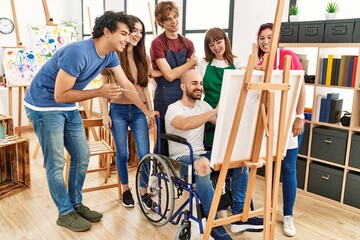 Group of young paint students smiling happy and looking draw of disabled partner sitting on wheelchair at art studio.
