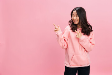 Lovely cheerful Asian student young lady in pink hoodie sweatshirt point finger aside at copy space for ad posing isolated on over pink studio background. Good offer. Fashion New Collection concept