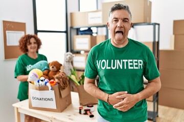 Middle age man wearing volunteer t shirt at donations stand sticking tongue out happy with funny...