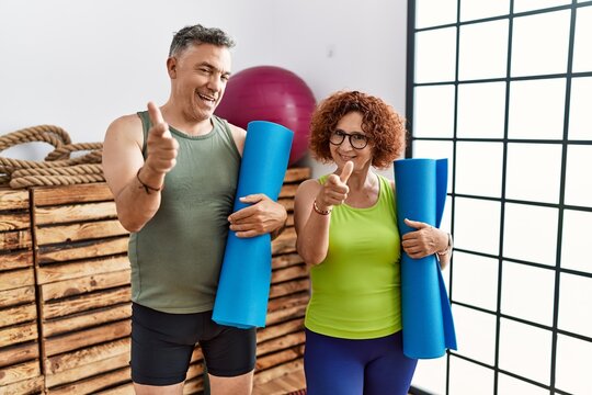 Middle age couple holding yoga mat pointing fingers to camera with happy and funny face. good energy and vibes. - Powered by Adobe