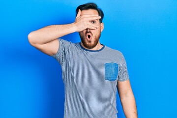 Handsome caucasian man with beard wearing casual striped t shirt peeking in shock covering face and eyes with hand, looking through fingers with embarrassed expression.