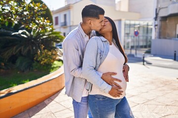 Young latin couple expecting baby touching belly and kissing at park