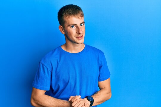 Young caucasian man wearing casual blue t shirt with hands together and crossed fingers smiling relaxed and cheerful. success and optimistic