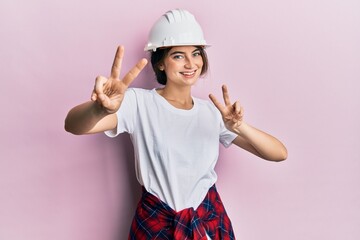Young caucasian woman wearing hardhat smiling looking to the camera showing fingers doing victory...