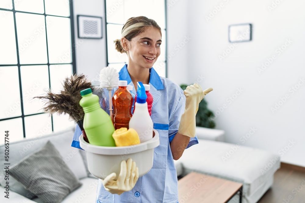 Canvas Prints Young blonde woman wearing cleaner uniform holding cleaning products pointing thumb up to the side smiling happy with open mouth