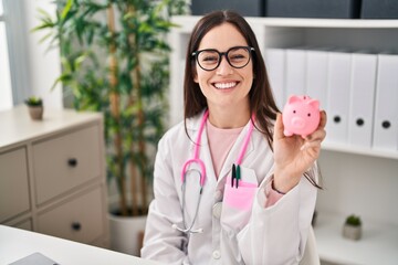 Young doctor woman holding piggy bank looking positive and happy standing and smiling with a...