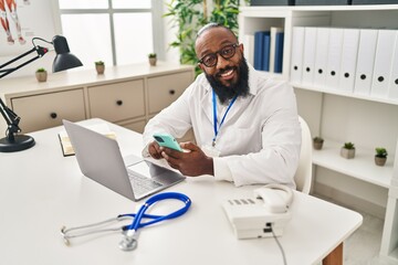 Naklejka na ściany i meble Young african american man wearing doctor uniform using smartphone working at clinic