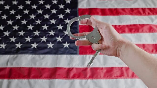 caucasian hand holding and turning silver metal handcuffs over blurry United States flag