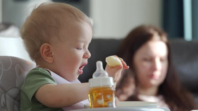 Baby boy with mother in the living room, cute kid sitting in booster seat and eating an apple, mom using smartphone. High quality 4k footage