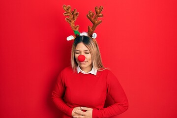 Beautiful hispanic woman wearing deer christmas hat and red nose with hand on stomach because indigestion, painful illness feeling unwell. ache concept.