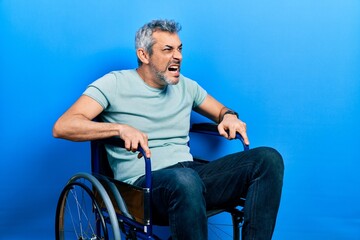 Handsome middle age man with grey hair sitting on wheelchair angry and mad screaming frustrated and furious, shouting with anger. rage and aggressive concept.