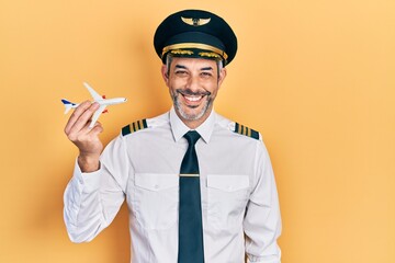 Handsome middle age man with grey hair wearing airplane pilot uniform holding toy plane looking positive and happy standing and smiling with a confident smile showing teeth © Krakenimages.com