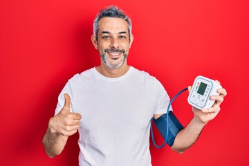 Handsome middle age man with grey hair using blood pressure monitor smiling happy and positive,...