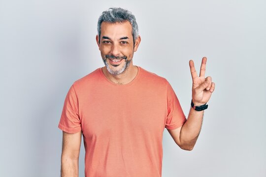 Handsome middle age man with grey hair wearing casual t shirt smiling with happy face winking at the camera doing victory sign. number two.