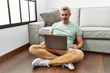Middle age hispanic man using laptop sitting on the floor at the living room skeptic and nervous,...