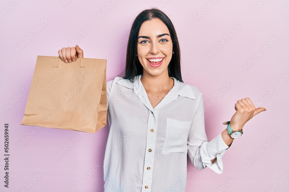 Poster beautiful woman with blue eyes holding take away paper bag pointing thumb up to the side smiling hap